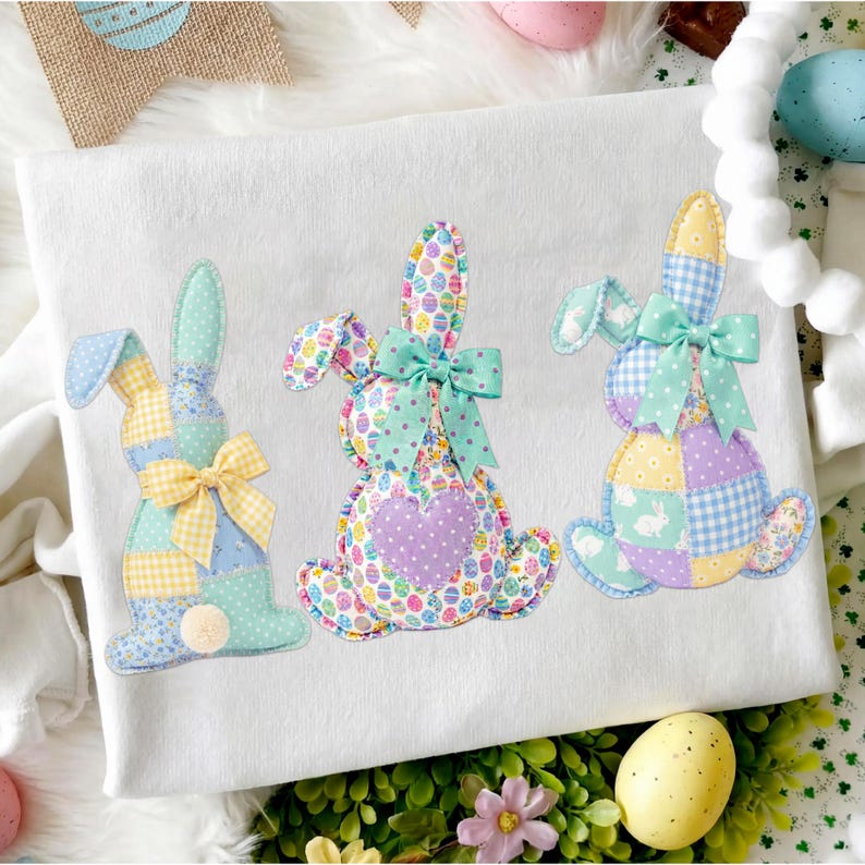 May include: White fabric with three appliqued Easter bunny designs. Each bunny is made of patchwork fabric in pastel colors, with bows and a pom-pom tail. The bunnies are decorated with egg and floral patterns.
