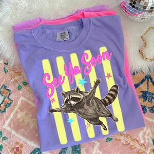 May include: A purple t-shirt with the phrase "See Ya Soon" in pink script, a raccoon graphic, and yellow vertical stripes. The shirt is folded on top of a pink t-shirt.