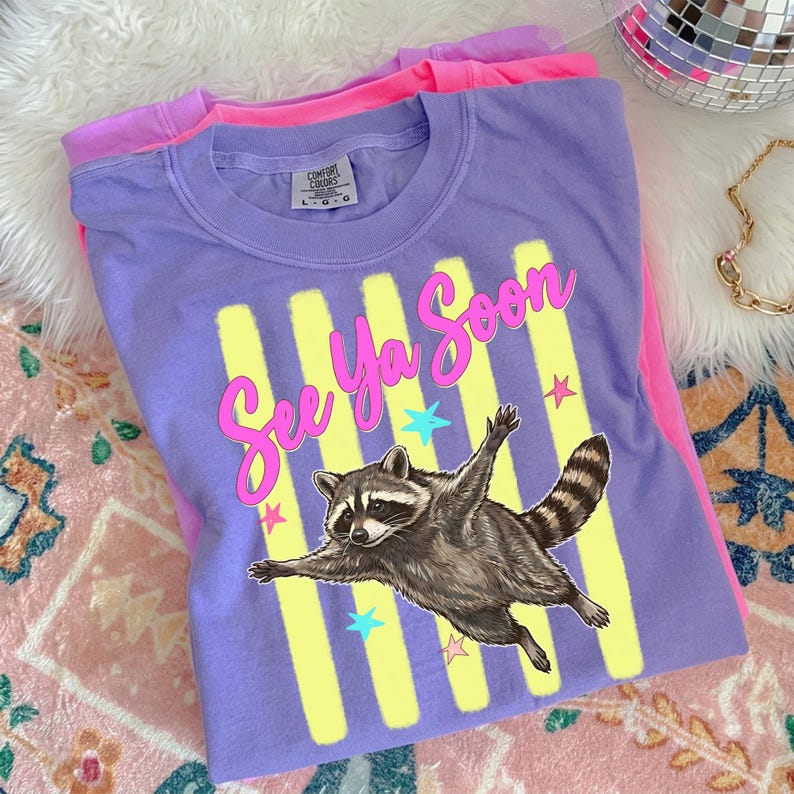May include: A purple t-shirt with the phrase "See Ya Soon" in pink script, a raccoon graphic, and yellow vertical stripes. The shirt is folded on top of a pink t-shirt.