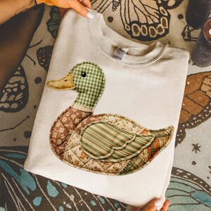 May include: Cream-colored sweatshirt featuring a patchwork duck design. The duck has a green gingham head, a brown floral body, and a green and orange striped wing. The design is quilted, adding texture to the garment.