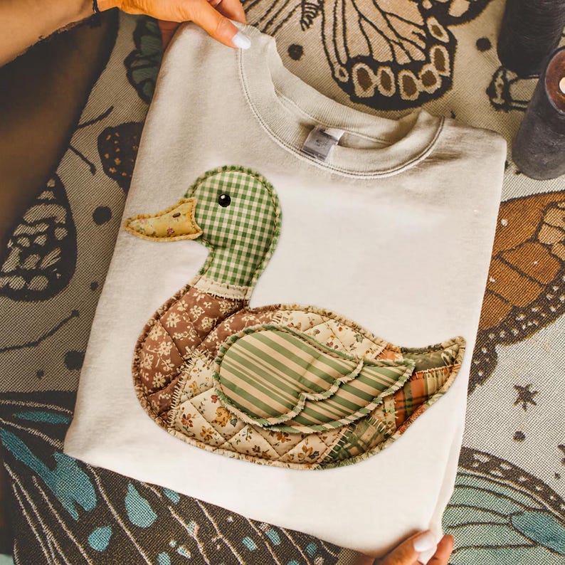 May include: Cream-colored sweatshirt featuring a patchwork duck design. The duck has a green gingham head, a brown floral body, and a green and orange striped wing. The design is quilted, adding texture to the garment.