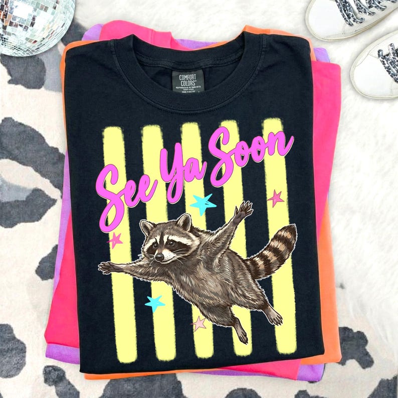 May include: Black t-shirt with a raccoon graphic and the text "See Ya Soon" in pink. The raccoon is brown and gray with its arms outstretched. The shirt is folded on top of pink, orange, and purple shirts.