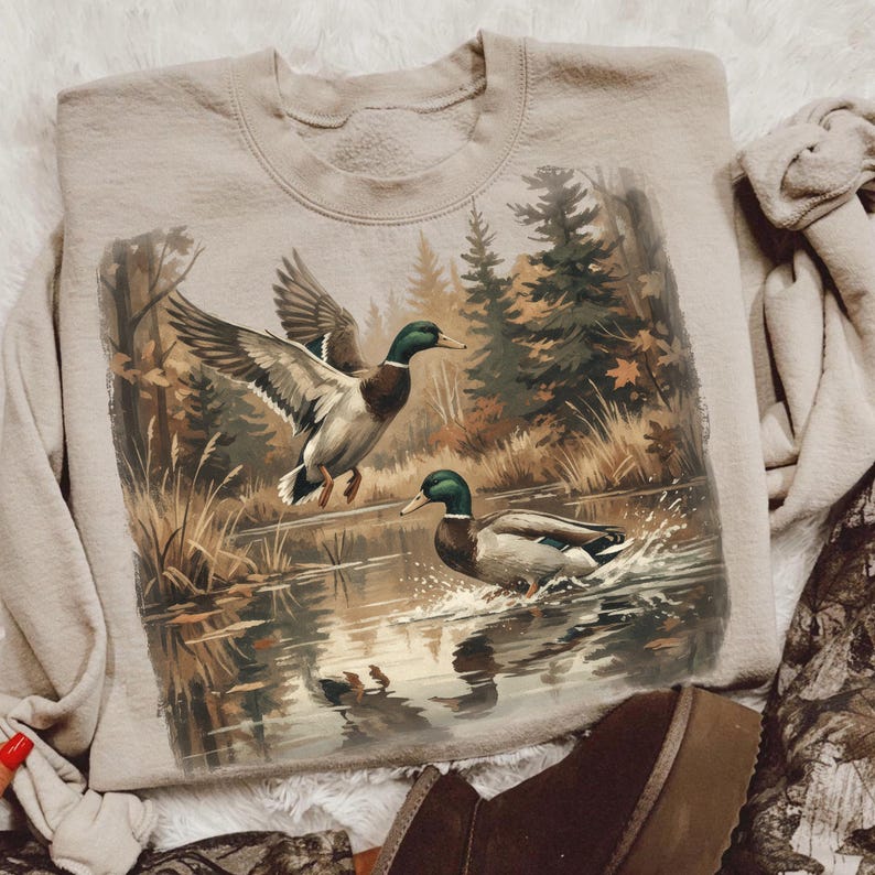 May include: Beige sweatshirt featuring a detailed illustration of two ducks in a natural pond setting. One duck is in flight, wings spread, while the other is in the water. The background shows a forest with trees and foliage.