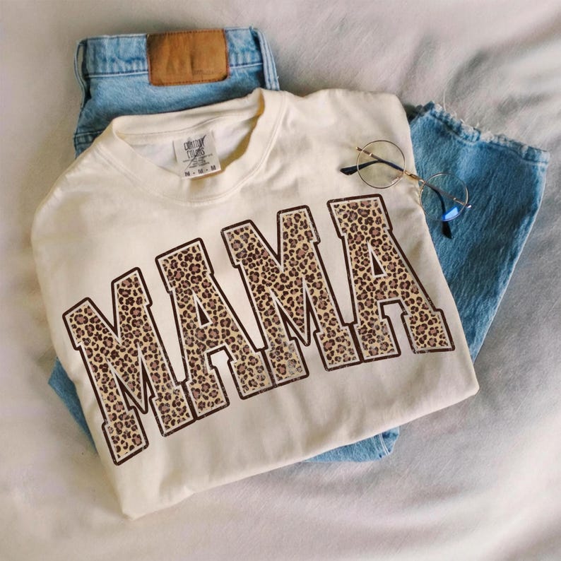 May include: Cream-colored t-shirt with the word "MAMA" in large, leopard print letters. The shirt is paired with light blue denim jeans and round-framed glasses. The shirt is a casual, everyday wear item.