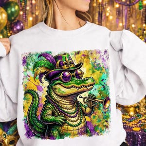 May include: White sweatshirt featuring a colorful illustration of an alligator playing a trumpet. The alligator wears a hat, sunglasses, and a beaded necklace, with a vibrant Mardi Gras-themed background. The design incorporates green, purple, and gold.