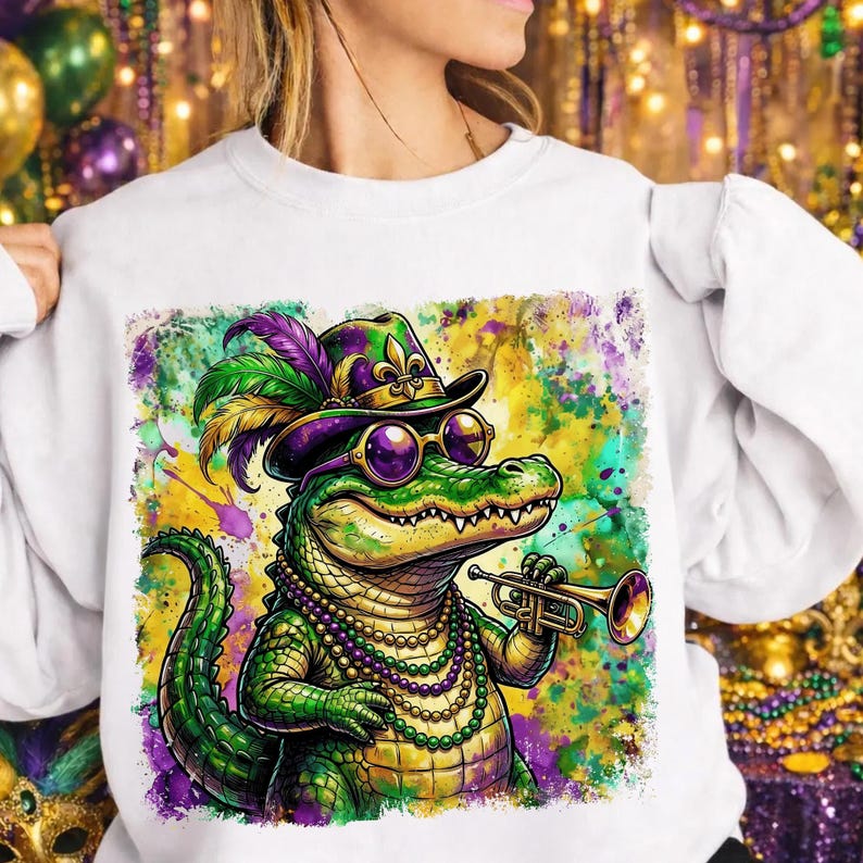 May include: White sweatshirt featuring a colorful illustration of an alligator playing a trumpet. The alligator wears a hat, sunglasses, and a beaded necklace, with a vibrant Mardi Gras-themed background. The design incorporates green, purple, and gold.