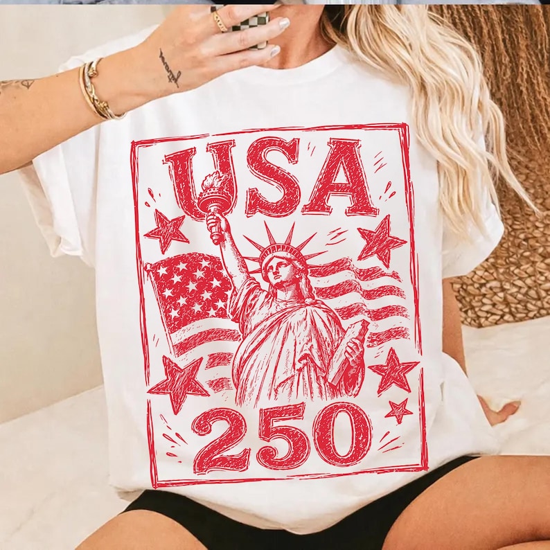May include: White t-shirt with a red graphic featuring the word "USA", the Statue of Liberty, a flag, stars, and the number "250". The design is enclosed in a red rectangular border.
