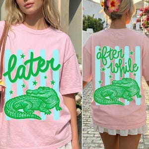 May include: Pink t-shirt with a green alligator graphic and the word "later" in green script on the front. The back of the shirt features the words "after while" and another alligator graphic. Light blue stripes and green stars accent the design.