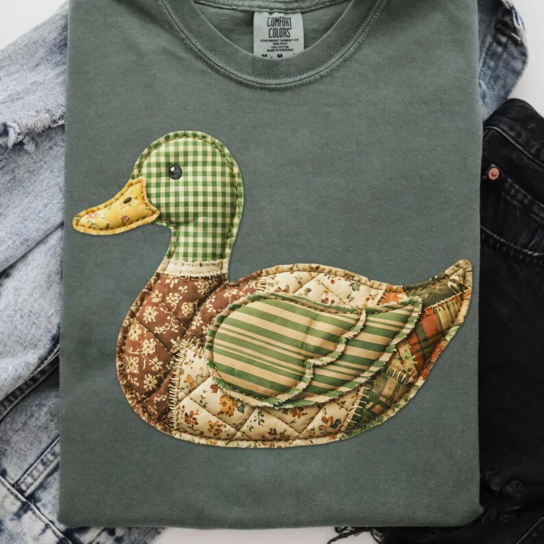 May include: Sage green t-shirt featuring a patchwork duck design. The duck has a green and white checkered head, a yellow beak, and a body made of various patterned fabrics in shades of brown, green, and cream. The shirt has the "Comfort Colors" brand label.