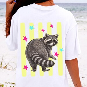May include: White t-shirt featuring a raccoon illustration with yellow vertical stripes and colorful stars. The raccoon is gray and black with a striped tail. The stars are pink and turquoise.