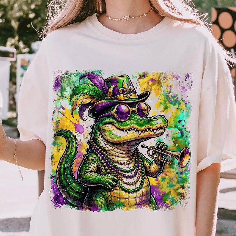May include: Cream-colored t-shirt featuring a vibrant illustration of a cartoon alligator. The alligator is wearing a purple and green hat, sunglasses, and a beaded necklace, holding a trumpet. The background has colorful paint splatters.