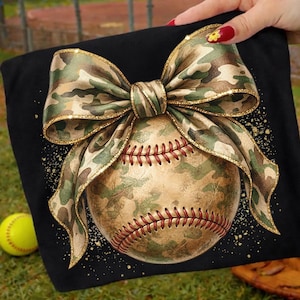 May include: Black t-shirt featuring a baseball with a camouflage pattern and red stitching. A large camouflage bow with gold trim is tied around the baseball. A yellow softball is in the background.