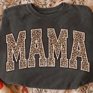May include: Dark gray sweatshirt with the word "MAMA" in large, bold letters. The letters are filled with a leopard print pattern and outlined in white. The sweatshirt is a casual, comfortable style.