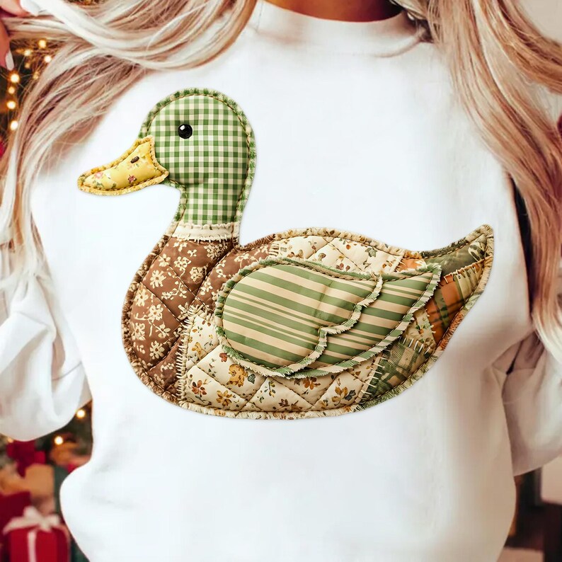 May include: A white sweatshirt featuring a patchwork duck design. The duck has a green and white checkered head, a yellow beak, and a body composed of various patterned fabrics in brown, green, and cream. The design is stitched onto the front of the sweatshirt.