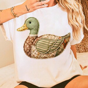 May include: White t-shirt featuring a patchwork duck design. The duck has a green and white checkered head, a yellow beak, and a body composed of various patterned fabrics in shades of brown, green, and cream. The design is centered on the shirt.