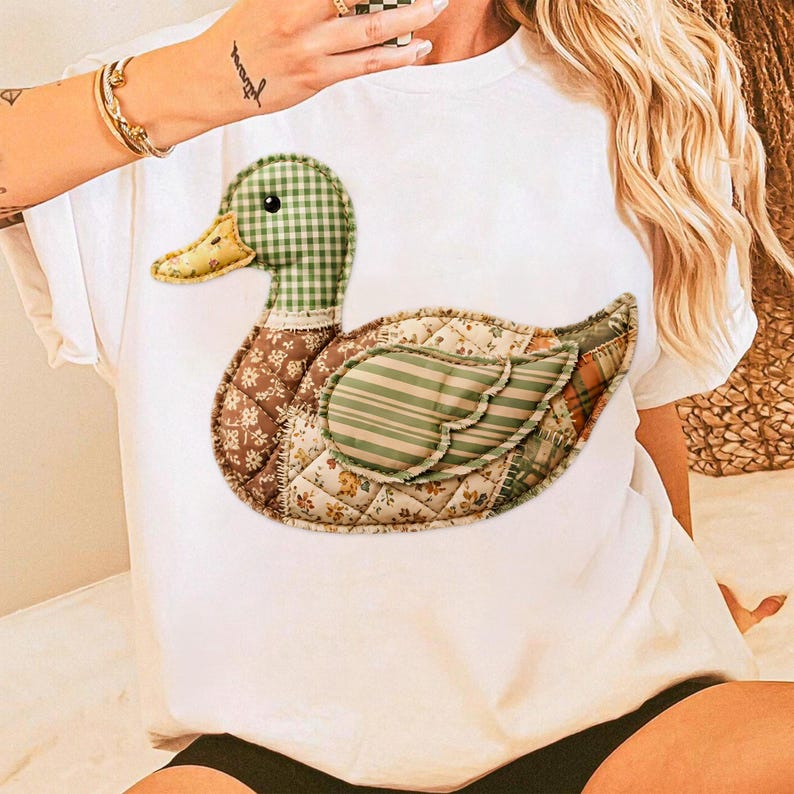 May include: White t-shirt featuring a patchwork duck design. The duck has a green and white checkered head, a yellow beak, and a body composed of various patterned fabrics in shades of brown, green, and cream. The design is centered on the shirt.