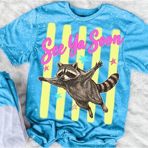 May include: A light blue t-shirt with a distressed, speckled design. The shirt features a raccoon illustration with outstretched arms, the text "See Ya Soon", yellow stripes, and star accents. The shirt is short-sleeved.