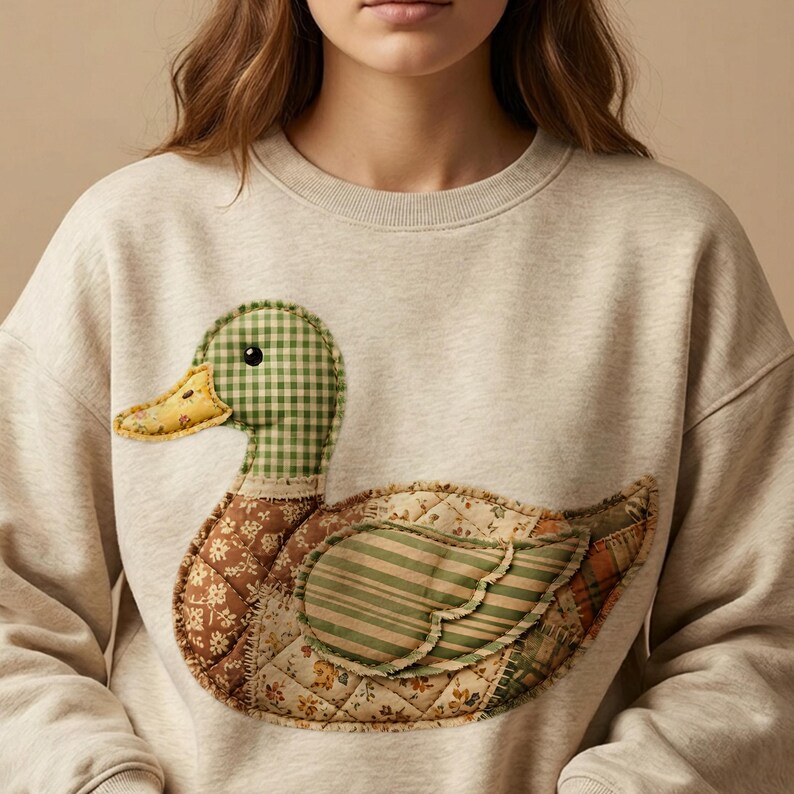May include: A light beige sweatshirt featuring a large, appliqu&eacute;d duck design. The duck has a green and white checkered head, a yellow beak, and a body composed of various patterned fabrics in brown, green, and yellow hues. The design is quilted.