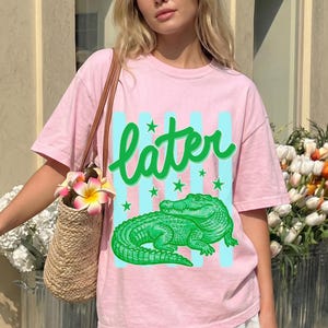 May include: A light pink t-shirt featuring a green alligator graphic with the word "later" in green script. The design includes light blue vertical stripes and green stars. The shirt is part of a casual wear collection.