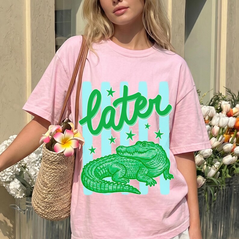 May include: A light pink t-shirt featuring a green alligator graphic with the word "later" in green script. The design includes light blue vertical stripes and green stars. The shirt is part of a casual wear collection.
