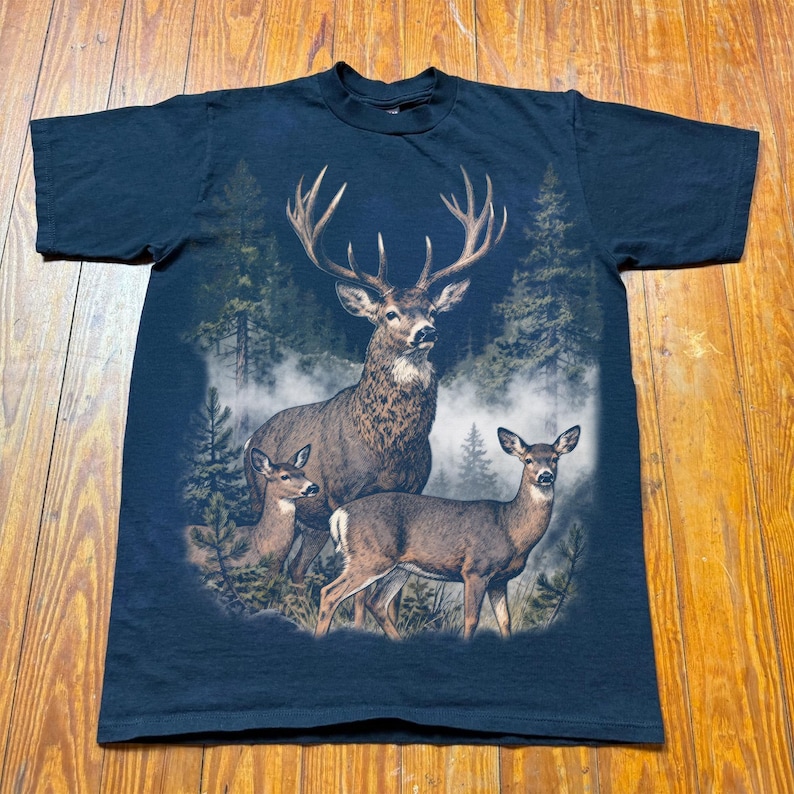 Peut inclure: T-shirt bleu marine avec un motif d&eacute;taill&eacute; d'un cerf avec de grands bois et deux biches dans un cadre forestier. Le motif comprend des arbres et un arri&egrave;re-plan brumeux, cr&eacute;ant une image sur le th&egrave;me de la nature.