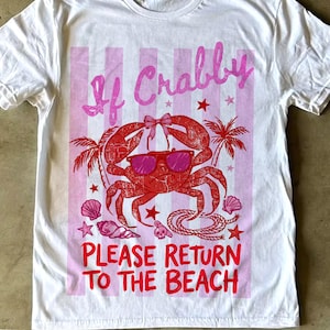 May include: White t-shirt with a pink and red graphic. The graphic features a red crab wearing sunglasses and a bow, with the text "If Crabby Please Return To The Beach". Palm trees, stars, and seashells are also included in the design.