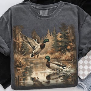 May include: Dark gray sweatshirt featuring a detailed illustration of two mallard ducks in a pond. One duck is in flight, wings spread, while the other is in the water, creating splashes. The background shows a forest scene with trees and reflections.