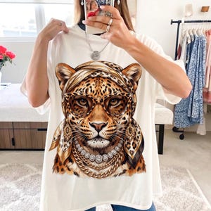 Op de afbeelding: Een wit t-shirt met een grafische print van een luipaard met een hoofddoek met luipaardprint, een bril met bruin montuur, gouden oorbellen, een diamanten ketting en een dikke gouden ketting.