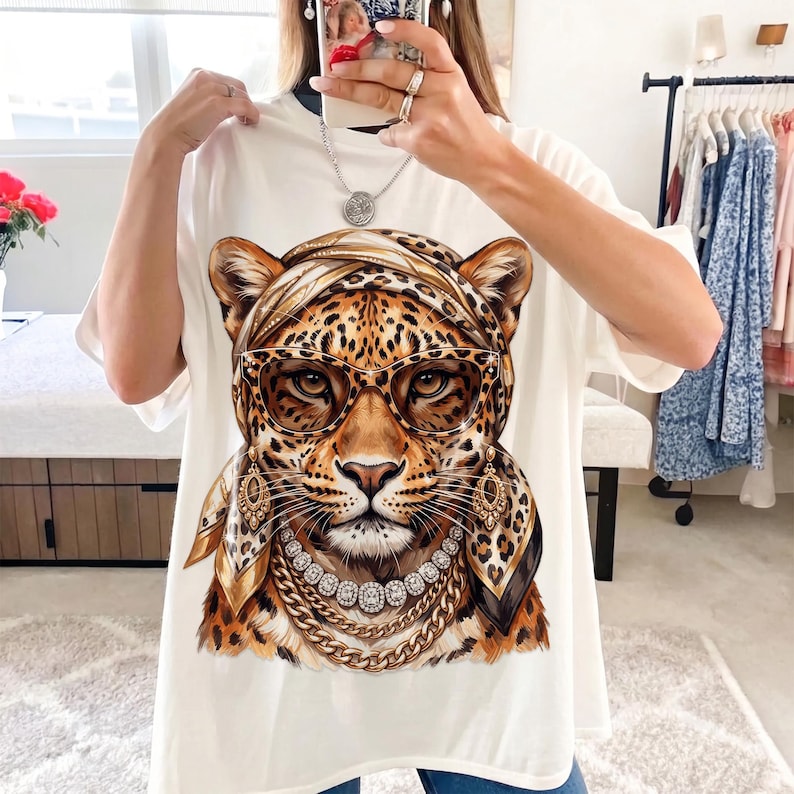 Op de afbeelding: Een wit t-shirt met een grafische print van een luipaard met een hoofddoek met luipaardprint, een bril met bruin montuur, gouden oorbellen, een diamanten ketting en een dikke gouden ketting.