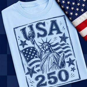 May include: Light blue t-shirt featuring a graphic of the Statue of Liberty holding a torch, with the word "USA" and the number "250" in a dark blue design. An American flag is draped in the background.