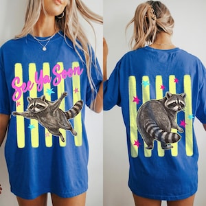 May include: Blue t-shirt with a raccoon graphic. The front features a raccoon with outstretched arms and the text "See Ya Soon" in pink. The back has a raccoon and vertical yellow stripes with stars.
