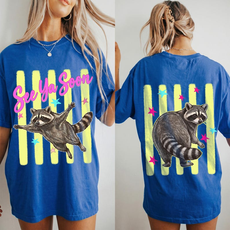 May include: Blue t-shirt with a raccoon graphic. The front features a raccoon with outstretched arms and the text "See Ya Soon" in pink. The back has a raccoon and vertical yellow stripes with stars.