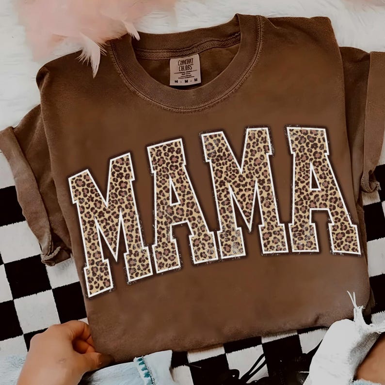 May include: A brown t-shirt with the word "MAMA" in large, block letters. The letters are filled with a leopard print pattern and outlined in white. The shirt has short sleeves and a crew neck.
