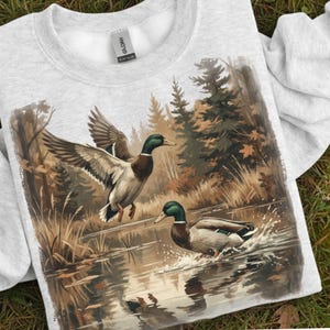 May include: A light gray sweatshirt featuring a detailed illustration of two mallard ducks in a wetland scene. One duck is in flight, wings spread, while the other is in the water. The artwork includes trees and foliage.