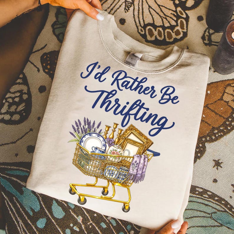 May include: Cream-colored sweatshirt with the text "I'd Rather Be Thrifting" in blue script. A shopping cart graphic is filled with vintage items like dishes, a picture frame, and lavender. The cart is yellow.