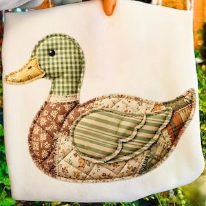 Op de afbeelding: Een gequilte eendenapplicatie op een witte achtergrond. De eend heeft een groen-witte geruite kop, een gele snavel en een lichaam met bloemen- en gestreepte stoffen. Het ontwerp heeft een landelijke uitstraling.