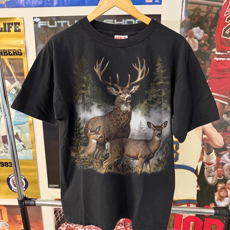 Peut inclure: T-shirt noir avec un motif d&eacute;taill&eacute; d'un cerf avec de grands bois et deux biches dans un cadre forestier. Le t-shirt a un col rond et des manches courtes. Le nom de la marque "WILD" est imprim&eacute; en haut du t-shirt.
