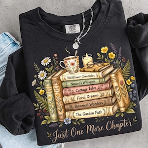 Puede incluir: Sudadera negra con un dise&ntilde;o de libros y flores. El dise&ntilde;o incluye una pila de libros con t&iacute;tulos como "Wildflower Chronicles" y una taza de t&eacute;. El texto "Just One More Chapter" est&aacute; en la parte inferior.