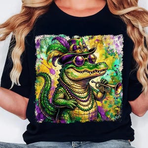 May include: Black t-shirt featuring a colorful illustration of an alligator wearing a hat, sunglasses, and a beaded necklace, playing a trumpet. The alligator is green and yellow, with purple and gold accents. The background is a splash of colors.