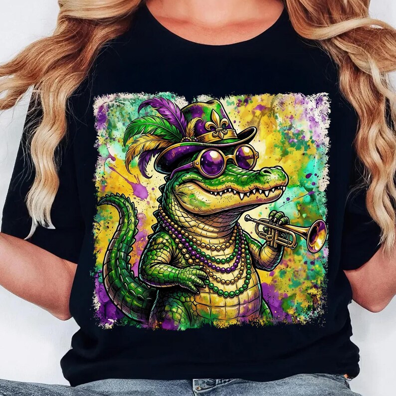 May include: Black t-shirt featuring a colorful illustration of an alligator wearing a hat, sunglasses, and a beaded necklace, playing a trumpet. The alligator is green and yellow, with purple and gold accents. The background is a splash of colors.