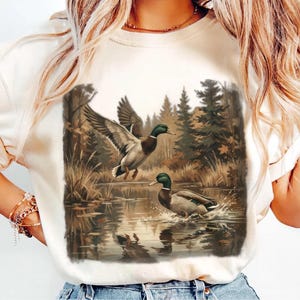 May include: Cream-colored sweatshirt featuring a detailed illustration of two ducks in a natural wetland setting. One duck is in flight, wings spread, while the other is in the water. The background includes trees and water reflections.