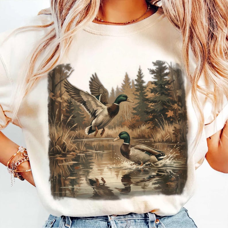 May include: Cream-colored sweatshirt featuring a detailed illustration of two ducks in a natural wetland setting. One duck is in flight, wings spread, while the other is in the water. The background includes trees and water reflections.