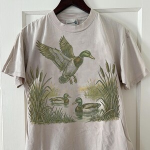 K&ouml;nnte beinhalten: Beiges Kurzarm-T-Shirt mit einem Naturmotiv mit Enten. Die Grafik zeigt eine Ente im Flug &uuml;ber zwei Enten im Wasser, mit Schilf und Rohrkolben. Das Shirt hat einen Rundhalsausschnitt und ein kleines Logo auf dem oberen R&uuml;cken.