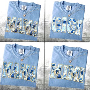 May include: Four light blue t-shirts, each embroidered with a different name in a patchwork design using blue and white floral and gingham fabrics. Each shirt features a gold necklace. The names displayed are "MAMA", "GIGI", "NANA", and "MIMI".