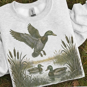 K&ouml;nnte beinhalten: Ein hellgraues Sweatshirt mit einer detaillierten Illustration von Enten in einem Teich. Eine Ente ist im Flug, die Fl&uuml;gel ausgebreitet, w&auml;hrend zwei andere auf dem Wasser schwimmen. Schilf und Rohrkolben umrahmen die Szene und schaffen ein Design zum Thema Natur.