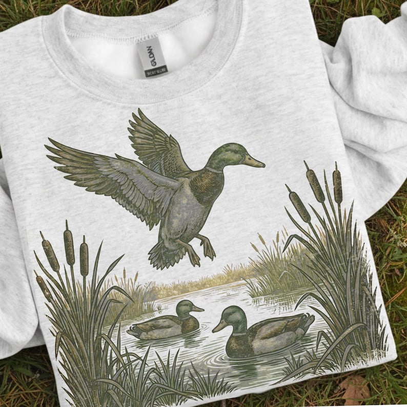 K&ouml;nnte beinhalten: Ein hellgraues Sweatshirt mit einer detaillierten Illustration von Enten in einem Teich. Eine Ente ist im Flug, die Fl&uuml;gel ausgebreitet, w&auml;hrend zwei andere auf dem Wasser schwimmen. Schilf und Rohrkolben umrahmen die Szene und schaffen ein Design zum Thema Natur.