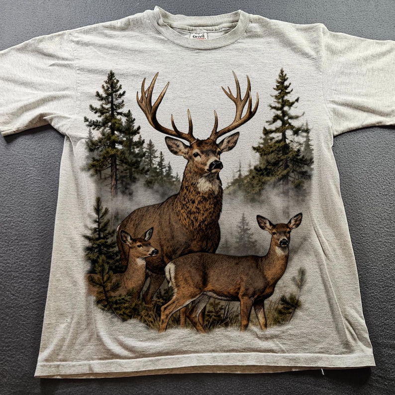 Peut inclure: Un t-shirt gris clair avec un motif d&eacute;taill&eacute; d'un cerf avec de grands bois, accompagn&eacute; de deux petits cerfs, sur fond de for&ecirc;t brumeuse avec des conif&egrave;res. Le t-shirt est fait d'une mati&egrave;re douce.