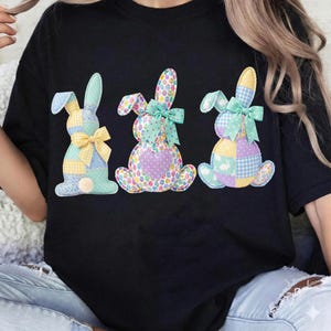 May include: Black t-shirt featuring three colorful patchwork bunny rabbits. Each bunny has a different pattern, including gingham, polka dots, and floral designs. The bunnies are adorned with bows and buttons, perfect for Easter.