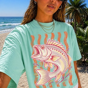 Könnte beinhalten: Hellblaues T-Shirt mit einer farbenfrohen Illustration eines Fisches mit offenem Maul. Der Fisch ist detailliert mit Gelb-, Rosa- und Rottönen. Vertikale orangefarbene Streifen und Wasserspritzer befinden sich hinter dem Fisch.