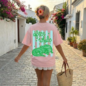 May include: A light pink t-shirt with the words "after while" in green script and a green alligator graphic. The shirt has a relaxed fit and is worn by a person walking down a cobblestone street.